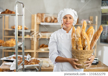 In working room of bakery, there is woman with baguettes in wicker basket in hands 131622270
