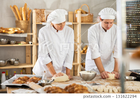 Two skilled female and male bakers in white uniform kneading dough and preparing portioned pieces before baking in bakehouse 131622294