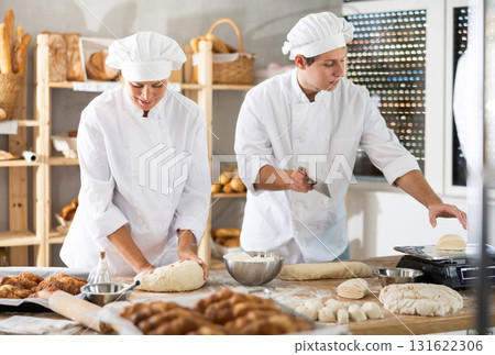 Female baker kneads dough while young guy cuts pieces of raw dough and weighs them on scales in bakery kitchen Female baker kneads dough while young guy cuts pieces of raw dough and weighs them on scales in bakery kitchen 131622306