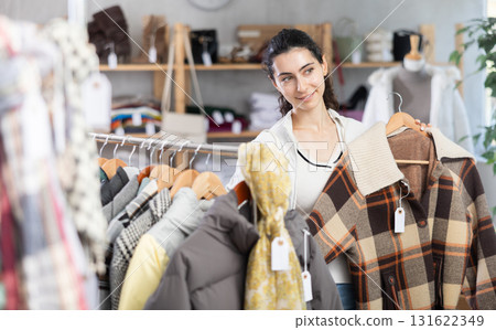 Female shopper chooses and try beautiful checkered coat-jacket in clothing store 131622349