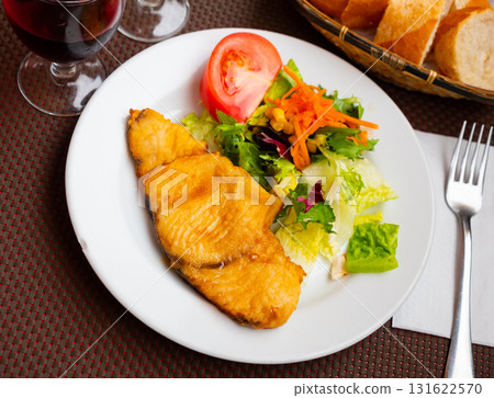 On plate there is small portion of salad of fresh vegetables and piece of fried fillet of white fish 131622570
