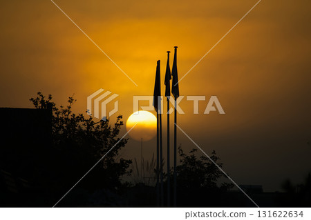 Silhouetted Flagpoles at Sunset: Warm Orange Sky with Rising Sun Behind Trees 131622634