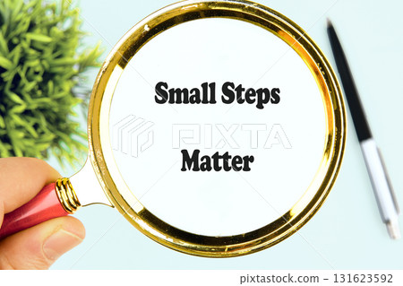 A hand holds a golden magnifying glass, focusing on the motivational message Small Steps Matter, a reminder of progress and achievement, placed near a pen and plant 131623592