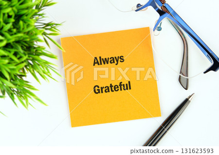 A yellow sticky note with the words Always Grateful sits on a desk, next to a pen, glasses, and a small potted plant, a reminder of positivity in business 131623593