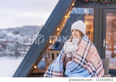 25- 35-year-old woman smiling and drinking hot tea, wrapped in a warm blanket, near her warm wooden triangular house on edge of a winter forest during the Christmas and New Year holidays in winter. 131628601