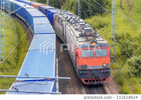 A red railway locomotive train with shipping containers rides next to a parallel train also with blue cargo containers among green hills close-up view from above. Railway transportation of goods 131628624