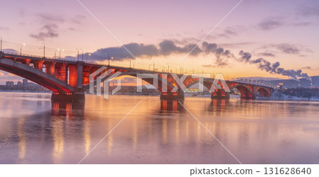 At dawn in Krasnoyarsk Russia, the Communal Bridge stands majestically over the Yenisei River. Its arches glow with vibrant hues as the sun rises, illuminating the tranquil waters below. 131628640