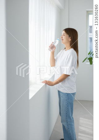 A Japanese woman holding a bottle of water while soaking up the natural light by the window 131630260