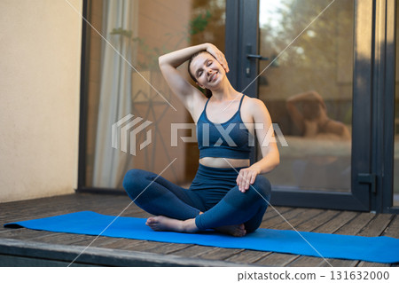 Young woman in sportswear sitting on yoga mat and doing neck stretching Young woman in sportswear sitting on yoga mat and doing neck stretching 131632000
