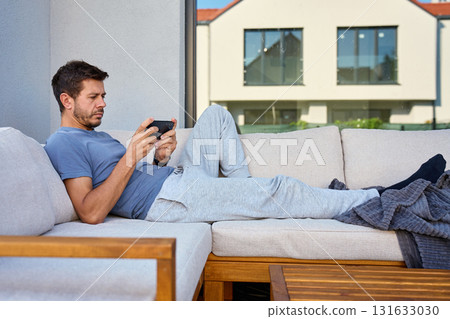 Man lying on sofa outdoors playing on smartphone Man lying on sofa outdoors playing on smartphone 131633030