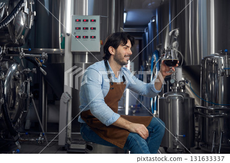 A man sits on a stool in a brewery holding a beer glass A man sits on a stool in a brewery holding a beer glass 131633337