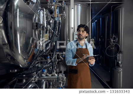 Brewer wearing an apron is holding a clipboard inside a brewery facility 131633343