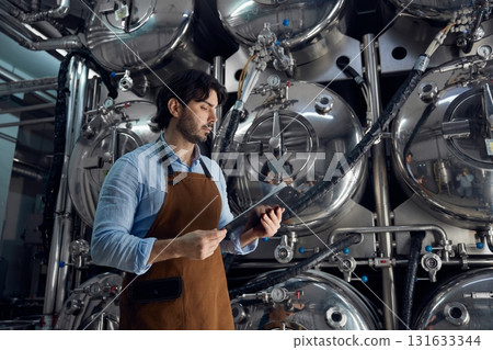 A man wearing an apron is holding a tablet inside a brewery facility A man wearing an apron is holding a tablet inside a brewery facility 131633344