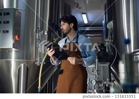 A man wearing an apron is utilizing a clipboard in a factory setting A man wearing an apron is utilizing a clipboard in a factory setting 131633351