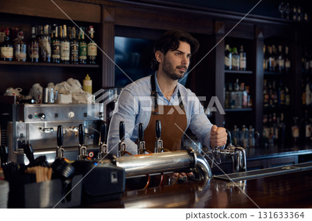 Handsome brewer is standing behind a bar counter, serving customers Handsome brewer is standing behind a bar counter, serving customers 131633364