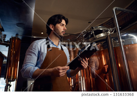 A man wearing an apron is studying a clipboard in a brewery setting 131633368