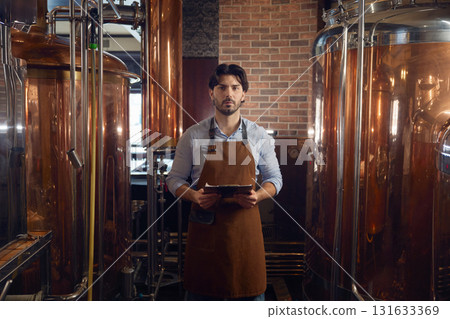 Confident man in an apron is holding a digital tablet inside a brewery Confident man in an apron is holding a digital tablet inside a brewery 131633369