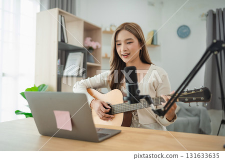 Young woman sings and plays guitar in a modern home studio, showcasing her talent during an online live performance with a laptop and microphone setup. 131633635