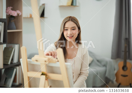 Young woman smiling while painting on canvas in cozy room with bright natural light, creative hobby, artistic expression, living space inspiration 131633638