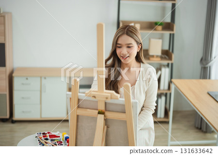 Young woman painting on an easel in a bright, modern studio filled with natural light, showcasing creativity and artistic expression in a serene environment. 131633642