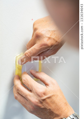 Man covering a light switch with masking tape before painting the wall white. Home renovation, surface protection and careful preparation during interior work. 131634643