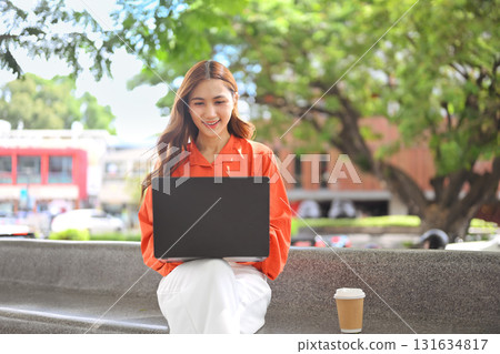 Smiling young woman sitting outside on a bench and using a laptop 131634817