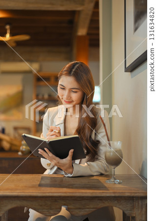 Smiling businesswoman in beige blazer sitting at a wooden table, taking notes in a notebook Smiling businesswoman in beige blazer sitting at a wooden table, taking notes in a notebook 131634820