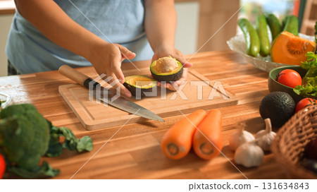 Hands cutting ripe avocado on wooden cutting board surrounded by fresh vegetables 131634843