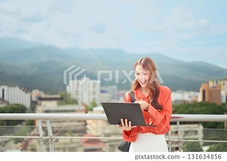 Smiling young woman using laptop outdoors on rooftop with city and mountain view Smiling young woman using laptop outdoors on rooftop with city and mountain view 131634856