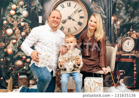 Parents and child pose near a decorated mantel with clock and festive ornaments. Natural composition, warm tones and gift boxes suitable for seasonal stock, lifestyle and retail imagery. 131635136