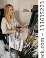 Woman working from home, sitting comfortably at bright white desk, typing on sleek white keyboard while smiling and engaging with computer screen in her cozy workspace. Professional blonde freelancer 131635223