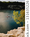 Stand up paddle boarding on calm lake with rocky cliff and trees in foreground and steep cliff with trees on opposite shore on sunny day. Paddleboarder gliding across tranquil lake, rocky shoreline 131635226