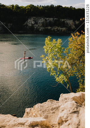Stand up paddle boarding on calm lake with rocky cliff and trees in foreground and steep cliff with trees on opposite shore on sunny day. Paddleboarder gliding across tranquil lake, rocky shoreline Stand up paddle boarding on calm lake with rocky cliff and trees in foreground and steep cliff with trees on opposite shore on sunny day. Paddleboarder gliding across tranquil lake, rocky shoreline 131635226