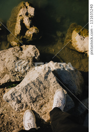 First-person view: person standing on large rocks by tranquil body of water, wearing white sneakers and black pants, soaking in the vibrant outdoor scenery on bright, sunny day. Men legs in sneakers First-person view: person standing on large rocks by tranquil body of water, wearing white sneakers and black pants, soaking in the vibrant outdoor scenery on bright, sunny day. Men legs in sneakers 131635240