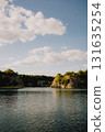 Calm lake water reflecting bright sky filled with fluffy white clouds, surrounded by rocky hills adorned with lush vegetation on sunny summer day, creating serene landscape. Tranquil lake. Nature 131635254