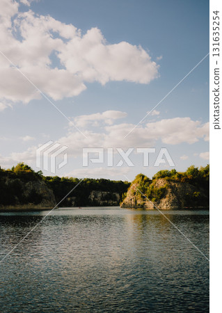 Calm lake water reflecting bright sky filled with fluffy white clouds, surrounded by rocky hills adorned with lush vegetation on sunny summer day, creating serene landscape. Tranquil lake. Nature 131635254