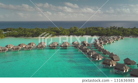 Aerial view luxury overwater bungalows resort nestled on pristine tropical island with turquoise water and lush green palm trees beach landscape. Exotic travel vacation honeymoon getaway. Drone flight 131636985