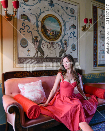 Woman in coral dress sitting on vintage sofa in ornate interior Woman in coral dress sitting on vintage sofa in ornate interior 131637147