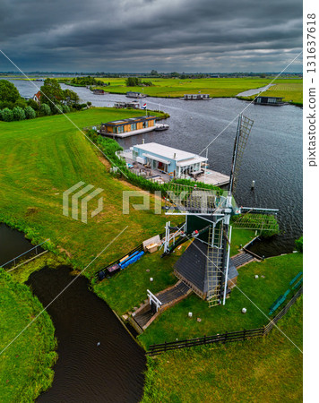 Aerial view of Dutch windmill beside canal with houseboats and modern buildings lush fields and flags add cultural texture beneath dramatic overcast skies. 131637618