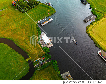 Aerial view of canal with moving boat, docks, windmill, and riverside buildings lush fields and water infrastructure blend tradition and recreation in a tranquil rural setting. 131637620