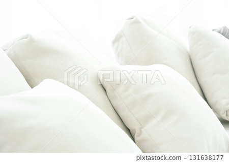 Closeup image of a pile of soft pillows made from white fabric used to decorate a sofa in a living room. There is natural light. It shines brightly from behind. 131638177