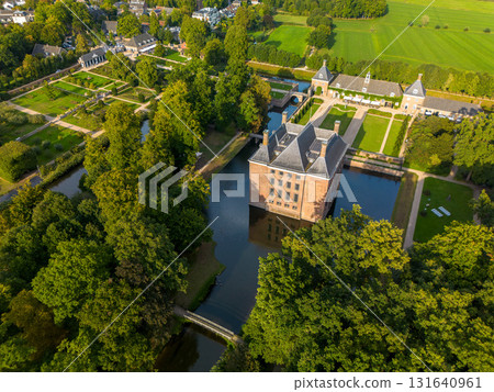 Aerial view of historic moated castle with gardens, bridge, and adjacent long building, surrounded by trees, water features, and a nearby village under warm sunlight. 131640961