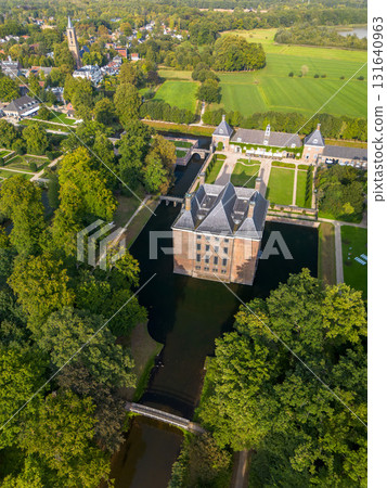 Aerial view of historic moated castle with gardens, bridge, and adjacent long building, surrounded by trees, water features, and a nearby village under warm sunlight. 131640963