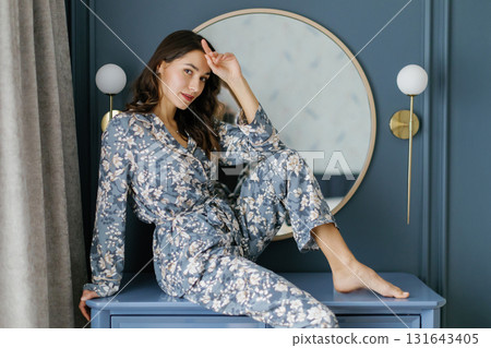 Woman wearing pajamas sitting on furniture in bedroom with round mirror and wall lamps Woman wearing pajamas sitting on furniture in bedroom with round mirror and wall lamps 131643405