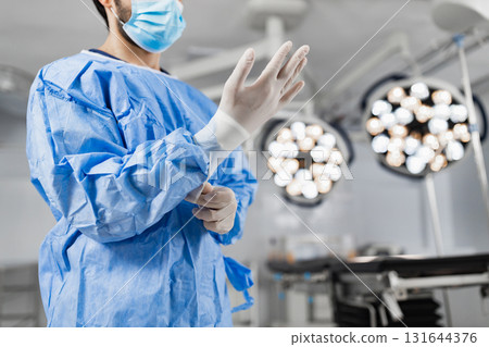 A surgeon in a blue surgical gown and mask puts on gloves in a bright operating room, preparing for a procedure. 131644376
