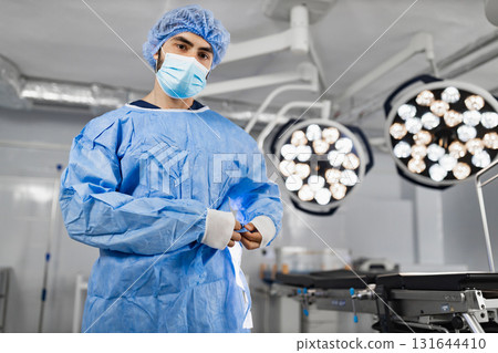 A surgeon in surgical attire stands in an operating room, ready for a procedure, with bright lights overhead. 131644410