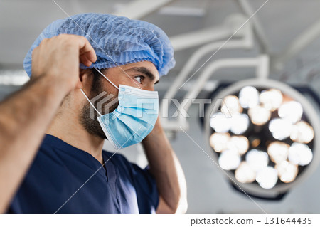 A surgeon adjusts his surgical cap and mask in a modern operating room, preparing for a procedure. 131644435
