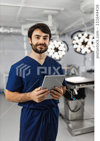 A surgeon stands in an operating room, holding a tablet and smiling at the camera. A surgeon stands in an operating room, holding a tablet and smiling at the camera. 131644438