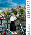 Fashionable woman poses by scenic canal, with bicycles and blooming flowers nearby, showcasing a relaxed urban atmosphere and vibrant lifestyle 131644807