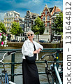 Fashionable woman poses by scenic canal, with bicycles and blooming flowers nearby, showcasing a relaxed urban atmosphere and vibrant lifestyle 131644812